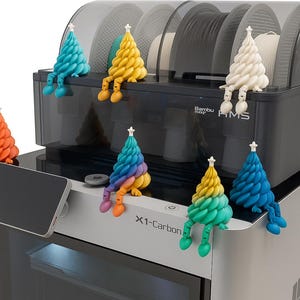 K&ouml;nnte beinhalten: Ein 3D-Drucker mit Touchscreen und mehreren bunten, dekorativen Weihnachtsbaumfiguren. Die B&auml;ume sind blau, gelb, orange, wei&szlig; und regenbogenfarben, jeweils mit einem Stern und Beinen, die &uuml;ber den Rand h&auml;ngen.