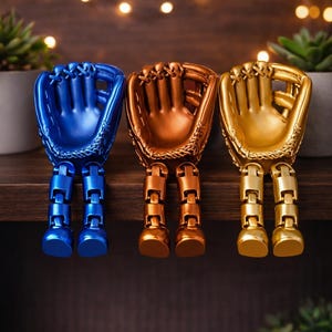 May include: Three baseball glove figurines in blue, bronze, and gold, each with articulated legs. The gloves are displayed on a wooden shelf with small potted plants and decorative lights in the background. The gloves are approximately 6 inches tall.