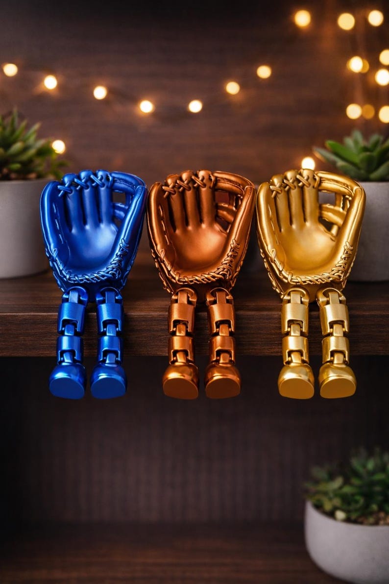 May include: Three baseball glove figurines in blue, bronze, and gold, each with articulated legs. The gloves are displayed on a wooden shelf with small potted plants and decorative lights in the background. The gloves are approximately 6 inches tall.