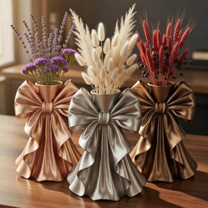 Puede incluir: Tres jarrones decorativos con dise&ntilde;o de lazo, cada uno con flores secas. Los jarrones son en tonos dorados, plateados y bronce. Las flores incluyen lavanda, blanco esponjoso y tallos rojos. Los jarrones est&aacute;n colocados sobre una superficie de madera.