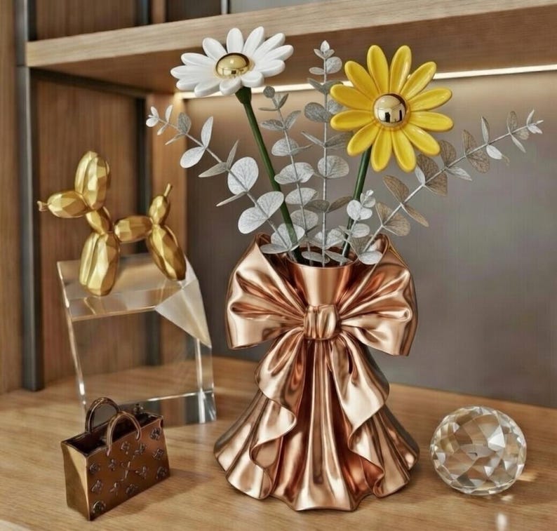 Puede incluir: Una composici&oacute;n decorativa con un jarr&oacute;n de oro rosa en forma de lazo, que contiene flores blancas y amarillas con forma de margarita con centros dorados. Tambi&eacute;n hay una escultura de perro dorado, un peque&ntilde;o bolso y una bola de cristal.