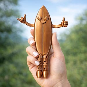 May include: A bronze surfboard-shaped ornament with a smiling face, arms, and legs. The figurine is held in a hand, with the arms in a "hang loose" and "thumbs up" gesture. The background is blurred green and blue.