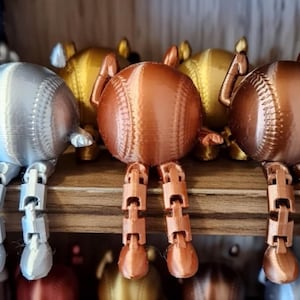 May include: A collection of baseball-shaped figurines in silver, gold, and copper colors. Each figurine has articulated legs, giving them a playful, whimsical appearance. The baseballs are detailed with stitching, and the legs are jointed.