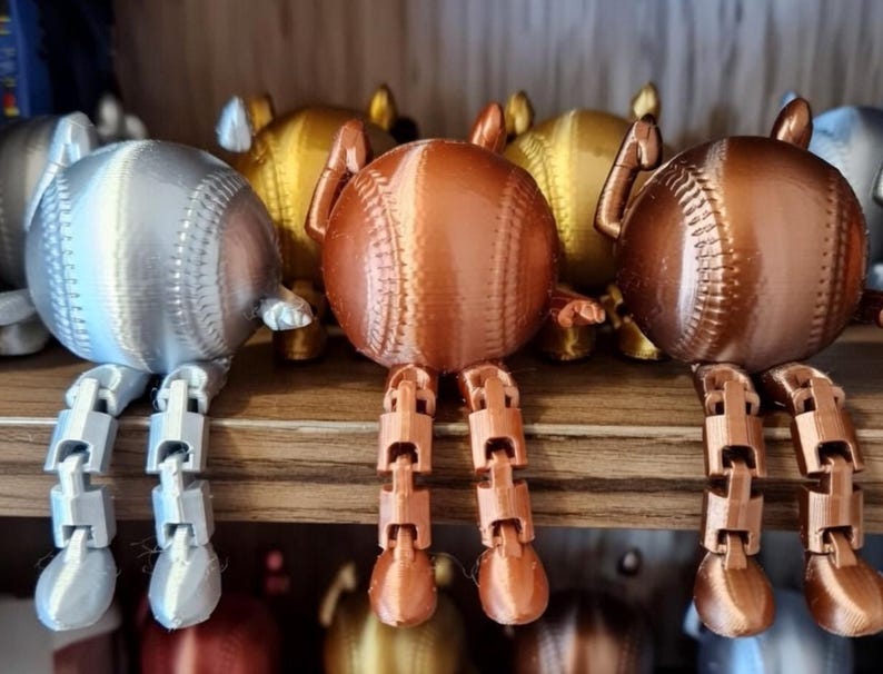 May include: A collection of baseball-shaped figurines in silver, gold, and copper colors. Each figurine has articulated legs, giving them a playful, whimsical appearance. The baseballs are detailed with stitching, and the legs are jointed.