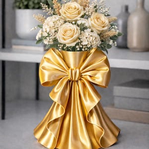 Puede incluir: Un jarr&oacute;n dorado con un gran dise&ntilde;o de lazo, lleno de un ramo de rosas color crema, flores blancas y espigas de trigo. El jarr&oacute;n tiene una base con volantes, creando un arreglo floral lujoso.