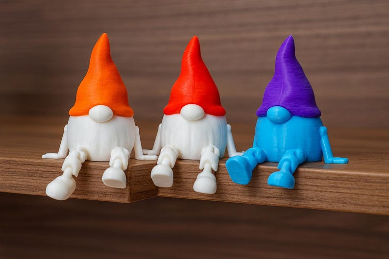 K&ouml;nnte beinhalten: Drei farbenfrohe Zwergfiguren sitzen auf einem Holzregal. Jeder Zwerg hat einen wei&szlig;en K&ouml;rper, eine runde wei&szlig;e Nase und einen farbigen Hut und Beine. Die H&uuml;te sind orange, rot und lila. Die Beine sind wei&szlig;, rot und blau.