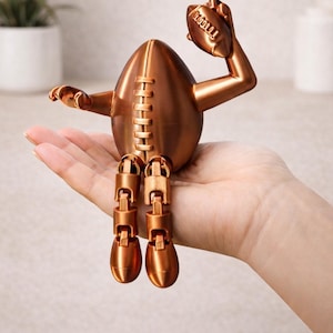 Puede incluir: Una escultura en forma de balón de fútbol americano, de color cobre, con piernas y brazos articulados, sosteniendo un balón de fútbol. La escultura tiene un detalle en forma de cremallera en el centro y está posada como si celebrara un touchdown.