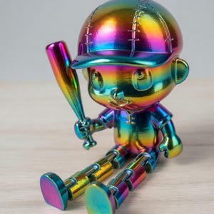 May include: A small, iridescent rainbow-colored figurine of a baseball player sitting down. The figure wears a cap and holds a baseball bat in one hand. The entire surface features a metallic, multi-colored gradient finish with visible joints at the knees.