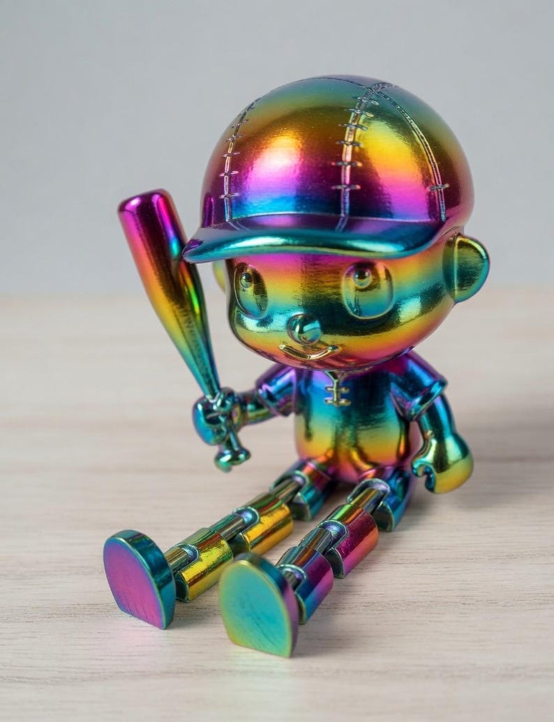 May include: A small, iridescent rainbow-colored figurine of a baseball player sitting down. The figure wears a cap and holds a baseball bat in one hand. The entire surface features a metallic, multi-colored gradient finish with visible joints at the knees.