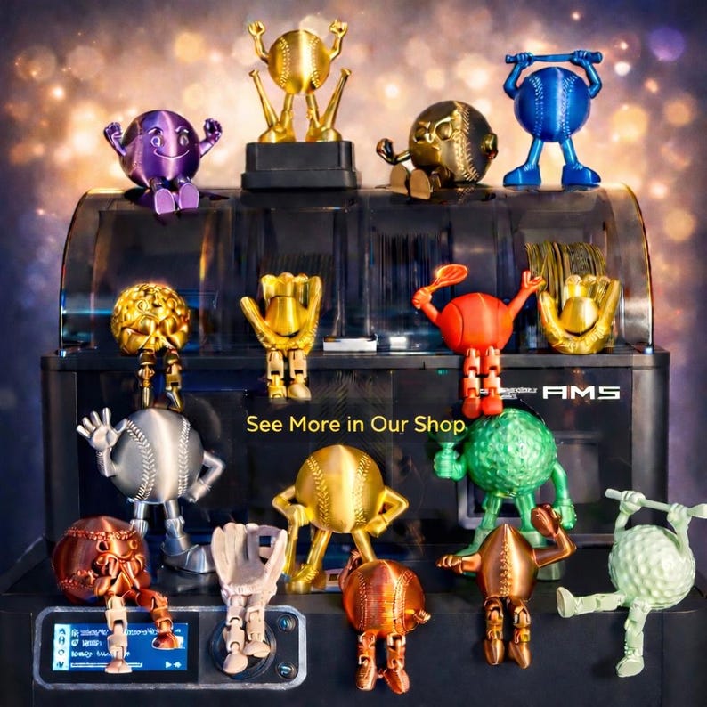 May include: A collection of colorful, 3D-printed sports-themed figurines, including baseballs, golf balls, and tennis balls with articulated limbs. The text "See More in Our Shop" is visible. The figurines are displayed on a black machine.