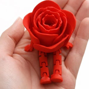 Puede incluir: Una figura de rosa roja vibrante con piernas articuladas y brazos pequeños. Los pétalos de la rosa son detallados y las piernas están hechas de segmentos conectados. La figura se sostiene en una mano sobre un fondo blanco.