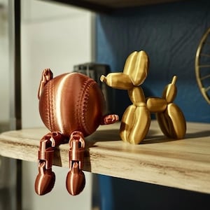 May include: A copper-colored baseball-shaped figurine with articulated legs sits next to a gold-colored balloon dog figurine. Both are displayed on a wooden shelf. The baseball figurine has visible stitching details.
