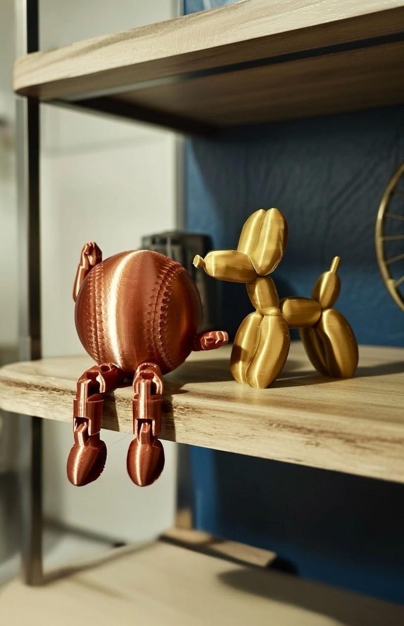 May include: A copper-colored baseball-shaped figurine with articulated legs sits next to a gold-colored balloon dog figurine. Both are displayed on a wooden shelf. The baseball figurine has visible stitching details.