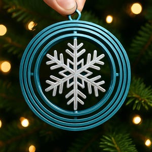 Könnte beinhalten: Ein türkisfarbenes und silbernes Schneeflocken-Weihnachtsornament. Das Ornament zeigt ein silbernes Schneeflocken-Design in der Mitte, umgeben von konzentrischen türkisfarbenen Ringen. Das Ornament wird von einem Finger und Daumen gehalten.