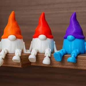 May include: Three colorful gnome figurines sitting on a wooden shelf. Each gnome has a white body, a round white nose, and a colored hat and legs. The hats are orange, red, and purple. The legs are white, red, and blue.