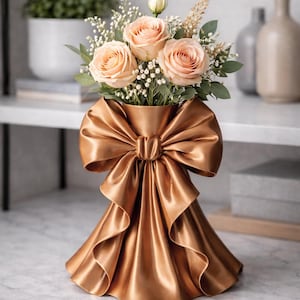 Puede incluir: Un jarrón dorado con diseño de lazo, que contiene un ramo de rosas melocotón y vegetación. El jarrón, con textura satinada, está sobre una superficie de mármol. El arreglo floral es elegante.