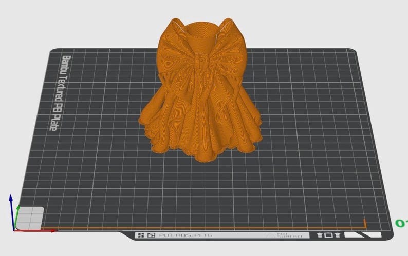 Puede incluir: Un lazo naranja impreso en 3D, con texturas y pliegues detallados, se encuentra sobre una superficie de cuadr&iacute;cula gris. El dise&ntilde;o del lazo es intrincado, con una apariencia en capas. El fondo incluye el texto "Bambu Textured PE Plate".