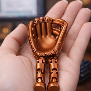 Puede incluir: Figura de guante de béisbol de color bronce con piernas articuladas. El guante presenta costuras detalladas y un diseño realista. Las piernas están compuestas por segmentos rectangulares y pies redondeados.