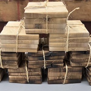 May include: Stacks of square wooden coasters, tied with twine. The coasters have a natural wood grain and a gradient of light to dark brown tones. The wood appears to be lightly charred, giving them a rustic look. The coasters are stacked in a pyramid shape.