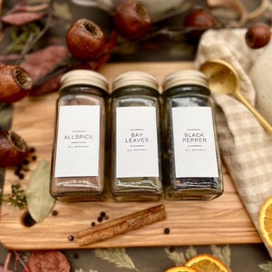K&ouml;nnte beinhalten: Drei Glas-Gew&uuml;rzgl&auml;ser mit wei&szlig;en Etiketten auf einem Holzbrett. Die Etiketten lauten "Allspice", "Bay Leaves" und "Black Pepper". Allspice ist dunkelbraun, Lorbeerbl&auml;tter sind gr&uuml;n und schwarzer Pfeffer ist schwarz. Die Gl&auml;ser stehen auf einem Holzbrett mit einem Zimtstange und schwarzen Pfefferk&ouml;rnern.