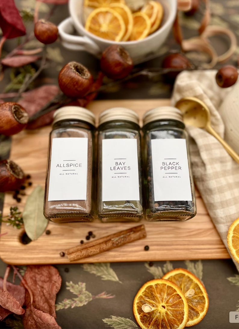 K&ouml;nnte beinhalten: Drei Glas-Gew&uuml;rzgl&auml;ser mit wei&szlig;en Etiketten auf einem Holzbrett. Die Etiketten lauten "Allspice", "Bay Leaves" und "Black Pepper". Allspice ist dunkelbraun, Lorbeerbl&auml;tter sind gr&uuml;n und schwarzer Pfeffer ist schwarz. Die Gl&auml;ser stehen auf einem Holzbrett mit einem Zimtstange und schwarzen Pfefferk&ouml;rnern.