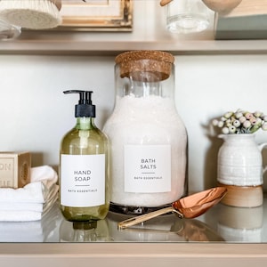 Peut inclure: Sur une étagère, une bouteille de savon pour les mains en verre transparent avec une pompe noire, un grand bocal de sels de bain et un petit vase blanc avec des fleurs. Une cuillère en cuivre et un savon sont également visibles. Les étiquettes indiquent "Savon pour les mains" et "Sels de bain".