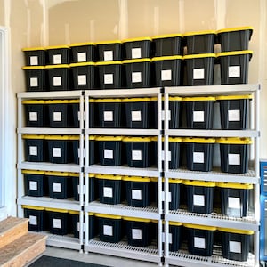 May include: A storage system with multiple black bins with yellow lids and white labels. The bins are stacked on white shelving units, creating an organized storage solution. The setting appears to be a garage or storage room.