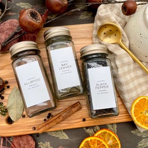 K&ouml;nnte beinhalten: Drei Glas-Gew&uuml;rzgl&auml;ser mit wei&szlig;en Etiketten und schwarzem Text. Die Etiketten lauten "Allspice", "Bay Leaves" und "Black Pepper". Die Gl&auml;ser stehen auf einer Holzoberfl&auml;che mit Zimtstangen, Lorbeerbl&auml;ttern und anderen Gew&uuml;rzen.