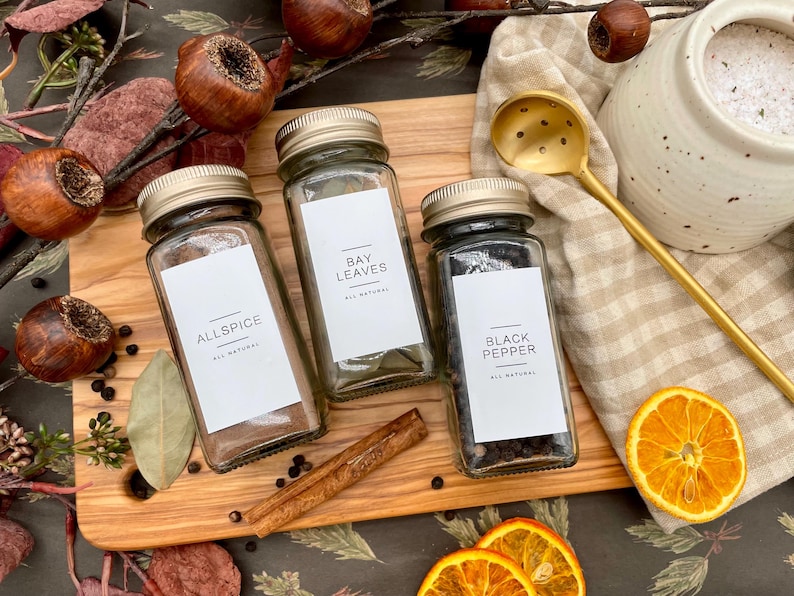 K&ouml;nnte beinhalten: Drei Glas-Gew&uuml;rzgl&auml;ser mit wei&szlig;en Etiketten und schwarzem Text. Die Etiketten lauten "Allspice", "Bay Leaves" und "Black Pepper". Die Gl&auml;ser stehen auf einer Holzoberfl&auml;che mit Zimtstangen, Lorbeerbl&auml;ttern und anderen Gew&uuml;rzen.