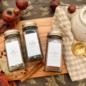 K&ouml;nnte beinhalten: Drei Glas-Gew&uuml;rzgl&auml;ser mit goldenen Deckeln und wei&szlig;en Etiketten auf einer Holzoberfl&auml;che. Die Etiketten lauten "Lorbeerbl&auml;tter", "Schwarzer Pfeffer" und "Piment". Alles nat&uuml;rlich.