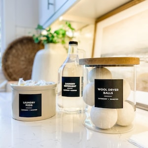May include: A laundry organisation set with a ceramic container labelled "Laundry Pods", a clear bottle of laundry detergent, and a glass jar of wool dryer balls. All items have black labels.