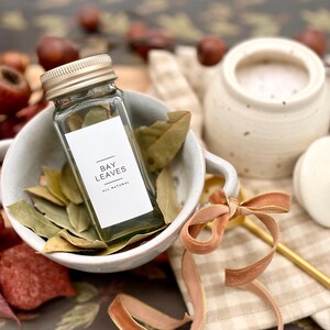 K&ouml;nnte beinhalten: Ein Glasgef&auml;&szlig; gef&uuml;llt mit getrockneten Lorbeerbl&auml;ttern, beschriftet "BAY LEAVES ALL NATURAL", steht in einer wei&szlig;en Sch&uuml;ssel mit weiteren Lorbeerbl&auml;ttern. Die Sch&uuml;ssel steht auf einem karierten Stoff mit einem goldenen L&ouml;ffel und einem braunen Band.