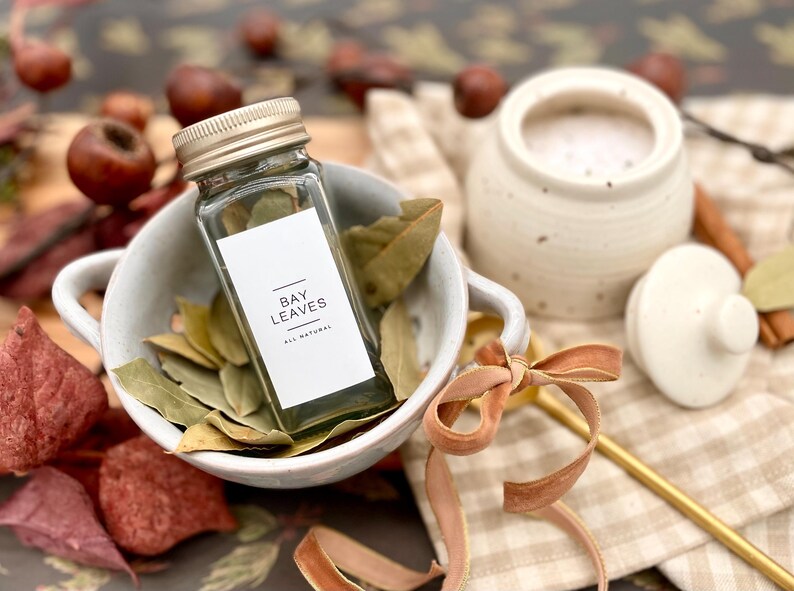 K&ouml;nnte beinhalten: Ein Glasgef&auml;&szlig; gef&uuml;llt mit getrockneten Lorbeerbl&auml;ttern, beschriftet "BAY LEAVES ALL NATURAL", steht in einer wei&szlig;en Sch&uuml;ssel mit weiteren Lorbeerbl&auml;ttern. Die Sch&uuml;ssel steht auf einem karierten Stoff mit einem goldenen L&ouml;ffel und einem braunen Band.