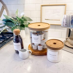 May include: A white countertop with three glass jars with wooden lids. The jars are labeled "Coffee Pods", "Creamer", and "Espresso".