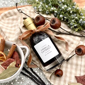 May include: A glass bottle of vanilla extract with a gold lid and a brown ribbon tied around the neck. The bottle is labeled "Vanilla Extract" with the words "All Natural" printed below. The bottle is sitting on a brown and white checkered fabric.