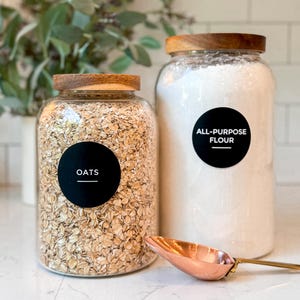 May include: Two glass jars with wooden lids, one filled with oats and the other with all-purpose flour. Black labels identify the contents. A copper scoop rests in front of the jars, suggesting a kitchen or pantry setting.