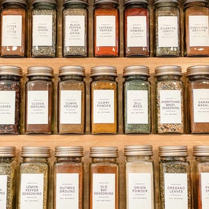 K&ouml;nnte beinhalten: Eine Nahaufnahme eines Gew&uuml;rzregals mit Glasbeh&auml;ltern, die mit verschiedenen Gew&uuml;rzen gef&uuml;llt sind. Die Beh&auml;lter sind mit den Namen der Gew&uuml;rze beschriftet, wie z. B. Apfelessig, Basilikumbl&auml;tter, schwarzer Pfeffer, Cayennepfeffer, Chilipulver, Korianderbl&auml;tter, Zimt gemahlen, Nelken gemahlen, Kreuzk&uuml;mmel gemahlen, Currypulver, Dillkraut, Alles-Bagel-Gew&uuml;rz, Garam Masala, Zitronenpfeffer-Gew&uuml;rz, Muskatnuss gemahlen, Old Bay Gew&uuml;rz, Zwiebelpulver, Oreganobl&auml;tter und Paprika.