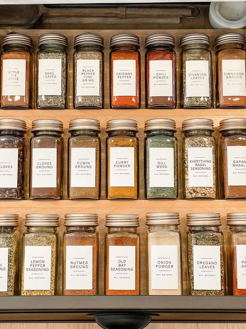 K&ouml;nnte beinhalten: Eine Nahaufnahme eines Gew&uuml;rzregals mit Glasbeh&auml;ltern, die mit verschiedenen Gew&uuml;rzen gef&uuml;llt sind. Die Beh&auml;lter sind mit den Namen der Gew&uuml;rze beschriftet, wie z. B. Apfelessig, Basilikumbl&auml;tter, schwarzer Pfeffer, Cayennepfeffer, Chilipulver, Korianderbl&auml;tter, Zimt gemahlen, Nelken gemahlen, Kreuzk&uuml;mmel gemahlen, Currypulver, Dillkraut, Alles-Bagel-Gew&uuml;rz, Garam Masala, Zitronenpfeffer-Gew&uuml;rz, Muskatnuss gemahlen, Old Bay Gew&uuml;rz, Zwiebelpulver, Oreganobl&auml;tter und Paprika.
