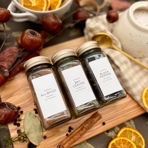 K&ouml;nnte beinhalten: Drei Glas-Gew&uuml;rzgl&auml;ser mit Etiketten mit der Aufschrift "Allspice", "Bay Leaves" und "Black Pepper". Die Gl&auml;ser stehen auf einem Holzbrett mit Zimtstangen und schwarzen Pfefferk&ouml;rnern.