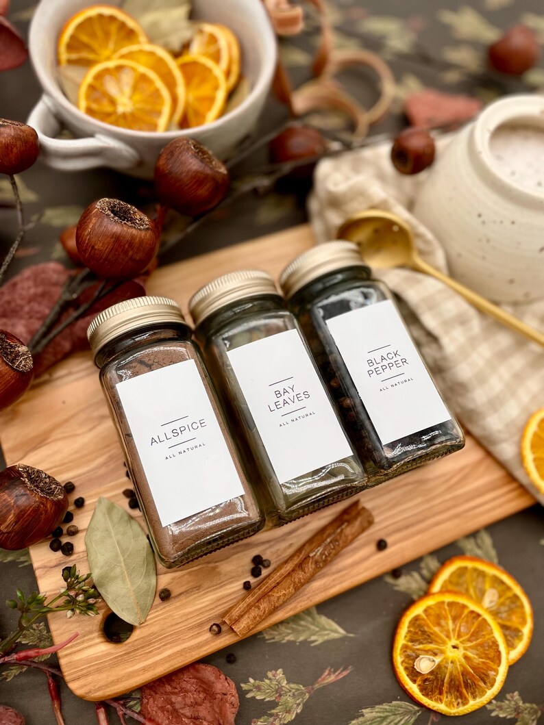 K&ouml;nnte beinhalten: Drei Glas-Gew&uuml;rzgl&auml;ser mit Etiketten mit der Aufschrift "Allspice", "Bay Leaves" und "Black Pepper". Die Gl&auml;ser stehen auf einem Holzbrett mit Zimtstangen und schwarzen Pfefferk&ouml;rnern.