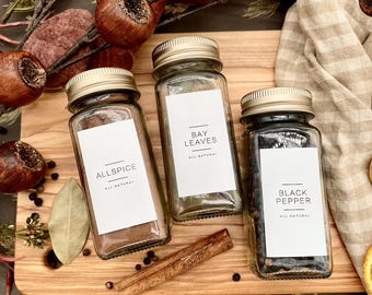 May include: Three glass jars with labels and gold lids filled with spices. The labels read "Allspice", "Bay Leaves", and "Black Pepper". Allspice is a dark brown color, bay leaves are green, and black pepper is black. The jars are on a wooden cutting board with a cinnamon stick.