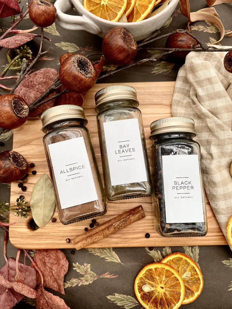K&ouml;nnte beinhalten: Drei Glasbeh&auml;lter mit Etiketten und goldenen Deckeln, gef&uuml;llt mit Gew&uuml;rzen. Die Etiketten lauten "Allspice", "Bay Leaves" und "Black Pepper". Allspice ist dunkelbraun, Lorbeerbl&auml;tter sind gr&uuml;n und schwarzer Pfeffer ist schwarz. Die Beh&auml;lter befinden sich auf einem Holzbrett mit einem Zimtstange.