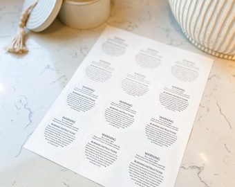 May include: A lit beige candle in a ceramic container with a lid and a sheet of white circular warning labels. The labels include the text "WARNING" and "BURNING INSTRUCTIONS". The candle is on a white surface.