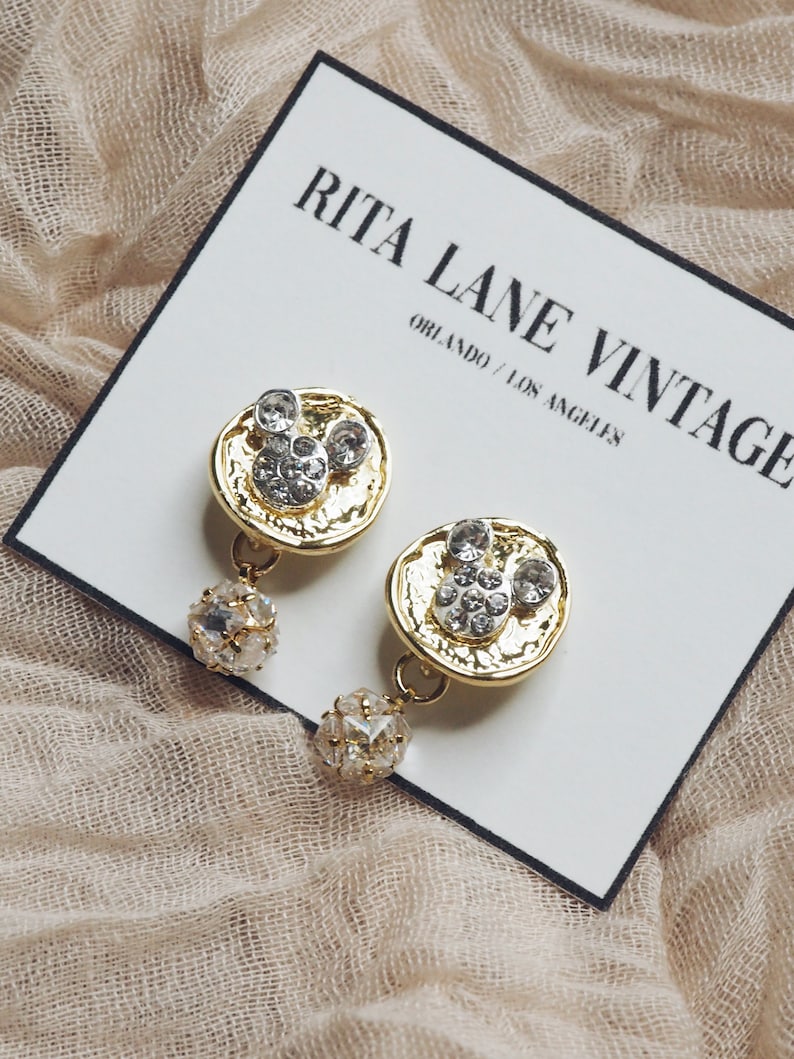 May include: Gold tone earrings with a Mickey Mouse design and clear rhinestones. The earrings have a dangling ball of clear rhinestones.