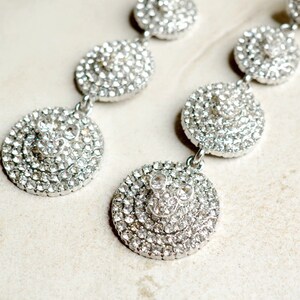 May include: A pair of silver dangle earrings with multiple layers of clear rhinestones. The earrings are circular and have a layered design.