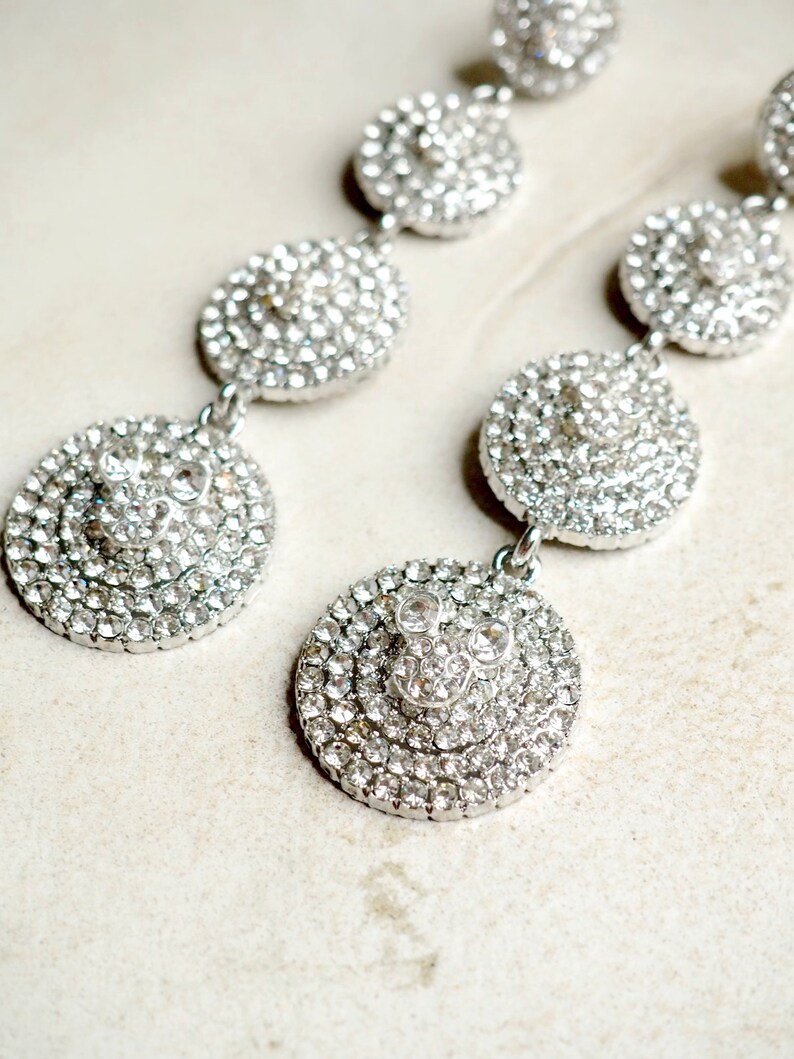 May include: A pair of silver dangle earrings with multiple layers of clear rhinestones. The earrings are circular and have a layered design.