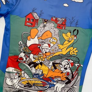 May include: A colorful cycling jersey featuring a cartoon design of Mickey Mouse, Donald Duck, Goofy, and Pluto riding bicycles. The jersey has a blue background with white clouds and green grass. The text "Hands!" is visible on the left side of the jersey.