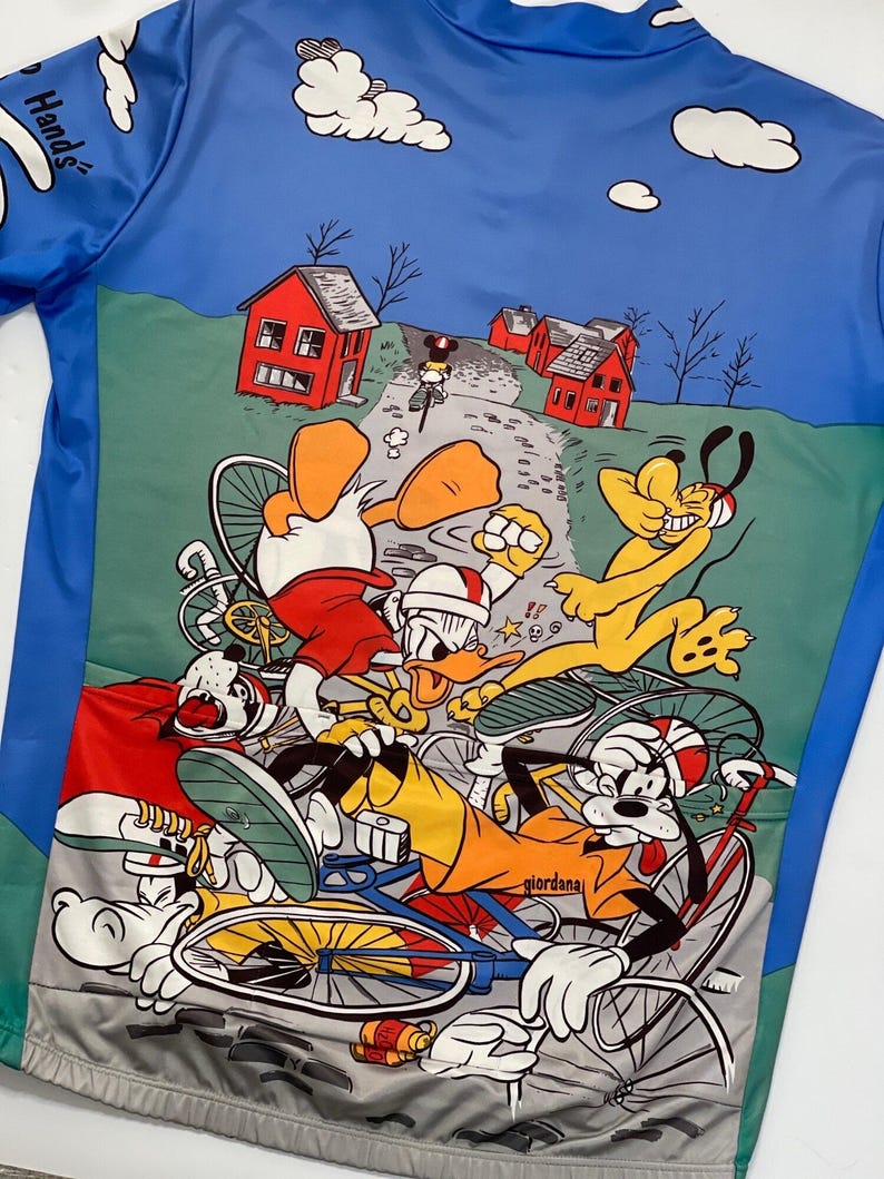 May include: A colorful cycling jersey featuring a cartoon design of Mickey Mouse, Donald Duck, Goofy, and Pluto riding bicycles. The jersey has a blue background with white clouds and green grass. The text "Hands!" is visible on the left side of the jersey.
