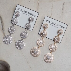 May include: Two pairs of dangle earrings with multiple round, crystal-encrusted charms. One pair is silver and the other is gold. The earrings are displayed on a white surface with a card that reads "Rita Lane Vintage" and "Old Radio / Los Angeles".