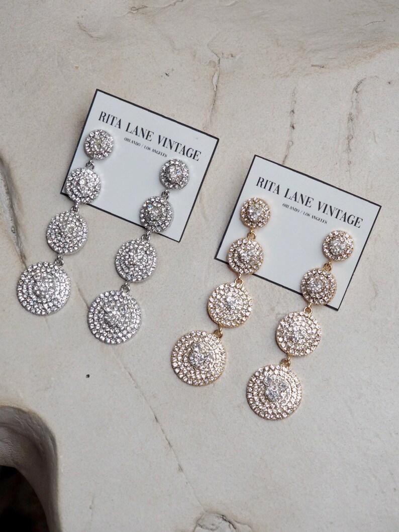 May include: Two pairs of dangle earrings with multiple round, crystal-encrusted charms. One pair is silver and the other is gold. The earrings are displayed on a white surface with a card that reads "Rita Lane Vintage" and "Old Radio / Los Angeles".